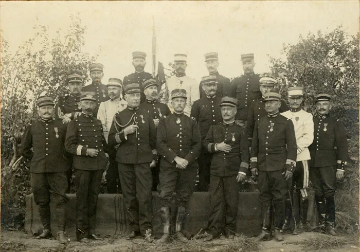 Photographie de groupe : une vingtaine d’officiers français posant en deux rangs devant un drapeau, en tenue de campagne et de service.