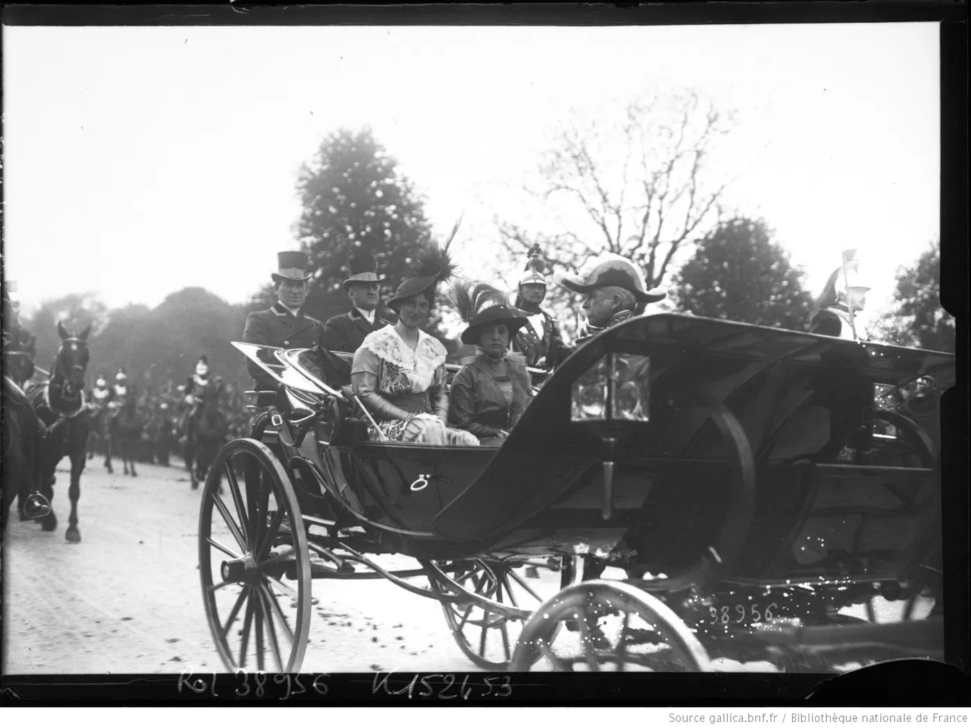 Plaque de verre : une calèche découverte transportant deux femmes en toilette et un officier en bicorne, escorte de cavalerie à l’arrière-plan.